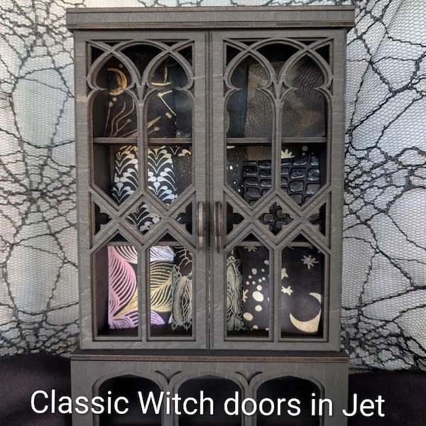 May include: A jet black wooden cabinet featuring two doors with intricate arched lattice cutouts and a lower shelf with three open compartments. The interior displays various patterned fabrics with celestial and botanical motifs. Text reads: Classic Witch doors in Jet Black.