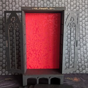 K&ouml;nnte beinhalten: Ein schwarzer Holz-Vitrinenschrank mit offenen T&uuml;ren, mit einem roten, gemusterten Innenraum. Der Schrank hat ein gotisches Bogendesign und enth&auml;lt mehrere schwarze Regale. Der Hintergrund ist ein Totenkopfmuster.