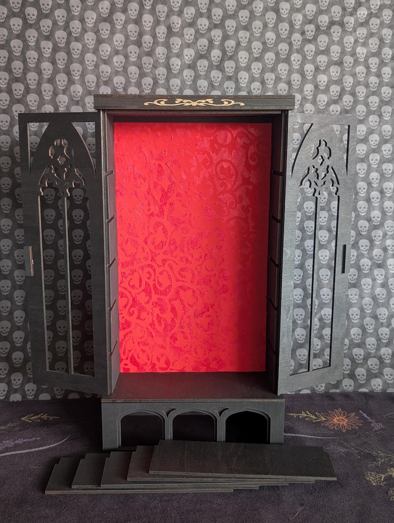 K&ouml;nnte beinhalten: Ein schwarzer Holz-Vitrinenschrank mit offenen T&uuml;ren, mit einem roten, gemusterten Innenraum. Der Schrank hat ein gotisches Bogendesign und enth&auml;lt mehrere schwarze Regale. Der Hintergrund ist ein Totenkopfmuster.