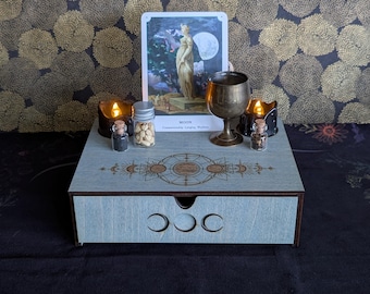 May include: A light blue wooden storage box with a drawer featuring three moon phase cutouts. The top surface displays a laser-engraved celestial design, a tarot card labeled MOON, a brass chalice, two small glass jars with corks, and two lit candles.