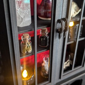 K&ouml;nnte beinhalten: Ein schwarzer Kuriosit&auml;tenschrank mit mehreren Regalen, der einen klaren Quarz-Kristall, eine kleine lila Vase und einen kleinen Sch&auml;del zeigt. Zwei kleine Glasflaschen mit Korken sind sichtbar, zusammen mit einer brennenden Kerze. Der Schrank hat einen dekorativen Griff.