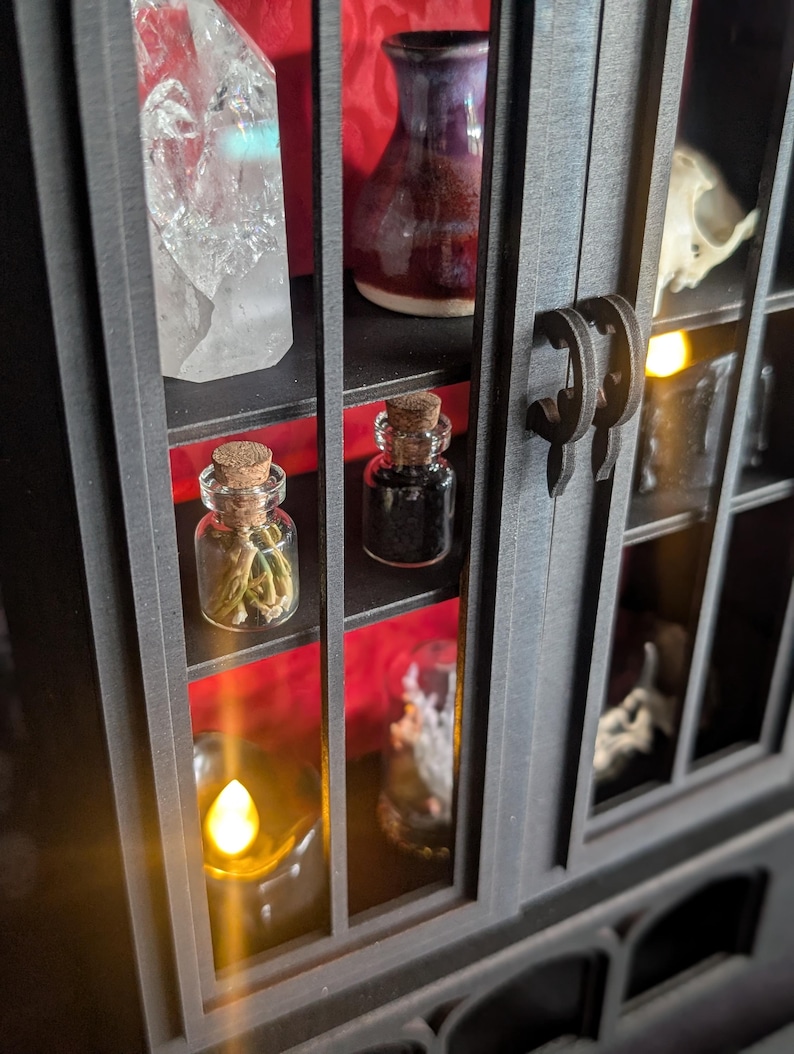K&ouml;nnte beinhalten: Ein schwarzer Kuriosit&auml;tenschrank mit mehreren Regalen, der einen klaren Quarz-Kristall, eine kleine lila Vase und einen kleinen Sch&auml;del zeigt. Zwei kleine Glasflaschen mit Korken sind sichtbar, zusammen mit einer brennenden Kerze. Der Schrank hat einen dekorativen Griff.