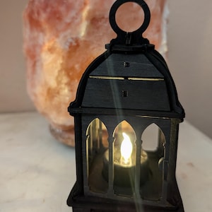 May include: A black wooden lantern with an arched window design and a handle. The lantern has a warm, flickering light inside, creating a cozy ambiance. The background features a large, pinkish-orange salt lamp.