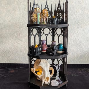 May include: A three-tiered black wooden corner shelf with gothic arch cutouts. The top shelf holds glass bottles and a black candle. The middle shelf features small ceramic vases and a black stone. The bottom shelf displays an animal skull.