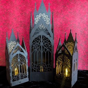 May include: Three black wooden gothic cathedral lanterns with intricate cut-out designs. The lanterns have a warm, inviting glow from the LED candles inside. The background is a textured red fabric, and the lanterns are on a black surface.