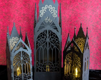 May include: Three black wooden gothic cathedral lanterns with intricate cut-out designs. The lanterns have a warm, inviting glow from the LED candles inside. The background is a textured red fabric, and the lanterns are on a black surface.