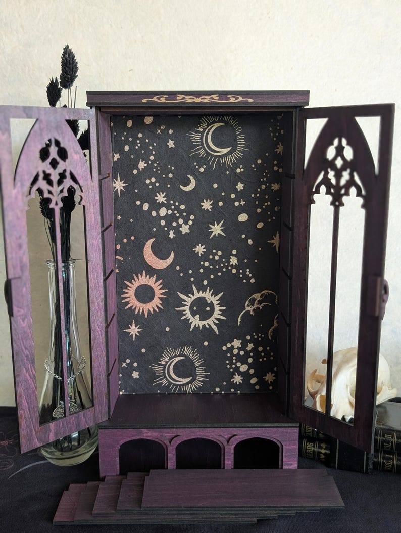 Puede incluir: Un gabinete de madera morada con dos puertas abiertas que presentan recortes en arco. El panel trasero interior es negro con patrones celestiales dorados que incluyen soles, lunas y estrellas. La base tiene tres aberturas en arco y escalones.