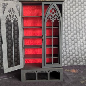 K&ouml;nnte beinhalten: Ein Miniatur-Schrank im gotischen Stil mit dunkelgrauer Au&szlig;enseite und leuchtend rotem Innenraum. Der Schrank hat ein bogenf&ouml;rmiges Fensterdesign und mehrere Regale. Die T&uuml;r ist ge&ouml;ffnet und gibt den Blick auf das Innere frei. Aufwendige Ausschnitte zieren das Design.