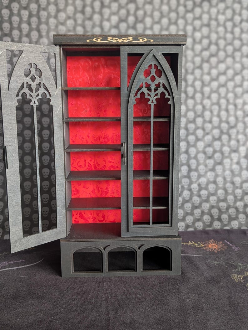 K&ouml;nnte beinhalten: Ein Miniatur-Schrank im gotischen Stil mit dunkelgrauer Au&szlig;enseite und leuchtend rotem Innenraum. Der Schrank hat ein bogenf&ouml;rmiges Fensterdesign und mehrere Regale. Die T&uuml;r ist ge&ouml;ffnet und gibt den Blick auf das Innere frei. Aufwendige Ausschnitte zieren das Design.