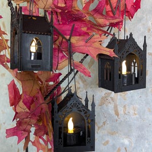 May include: Three black hanging ornaments shaped like miniature gothic buildings with pointed arches and spires. Each structure contains a small, battery-operated tea light with a warm yellow glow, suspended by thin black strings among red leaves.