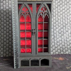 K&ouml;nnte beinhalten: Ein schwarzer Holzschrank mit gotischem Bogen-Design. Der Schrank hat zwei T&uuml;ren mit aufwendigen Ausschnitten und rotem Samtr&uuml;cken. Der Schrank ist etwa 30 cm hoch und hat Regale im Inneren.