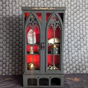 K&ouml;nnte beinhalten: Ein schwarzer Miniaturschrank mit gotischen Bogen-T&uuml;ren und Regalen, der einen Kristall, einen Sch&auml;del, Kerzen und Fl&auml;schchen vor rotem Hintergrund zeigt. Der Schrank hat goldene Akzente und steht auf einer dunklen Oberfl&auml;che.