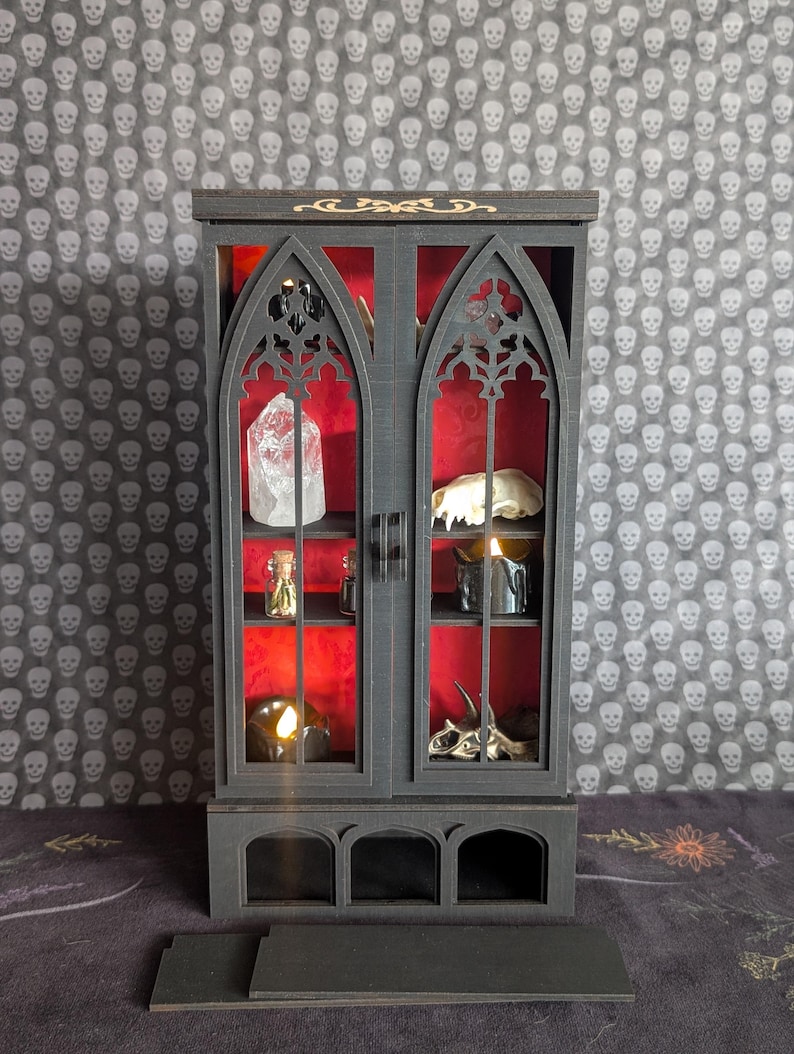 K&ouml;nnte beinhalten: Ein schwarzer Miniaturschrank mit gotischen Bogen-T&uuml;ren und Regalen, der einen Kristall, einen Sch&auml;del, Kerzen und Fl&auml;schchen vor rotem Hintergrund zeigt. Der Schrank hat goldene Akzente und steht auf einer dunklen Oberfl&auml;che.