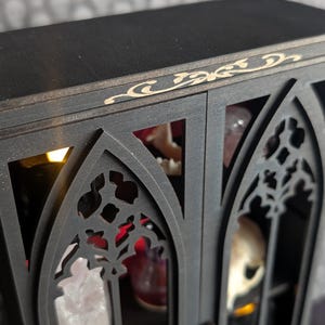 K&ouml;nnte beinhalten: Ein schwarzer Holzschrank mit gotischen Bogen-Ausschnitten und dekorativen Zierleisten. Der Schrank hat eine Innenbeleuchtung und ist mit verschiedenen Objekten gef&uuml;llt, darunter Kristalle und ein Sch&auml;del.