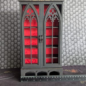 K&ouml;nnte beinhalten: Ein schwarzer Holzschrank mit zwei T&uuml;ren, der gotische Bogenfenster-Designs aufweist. Der Innenraum ist mit rotem Stoff ausgekleidet. Der Schrank hat Regale und ist etwa 25 cm breit.