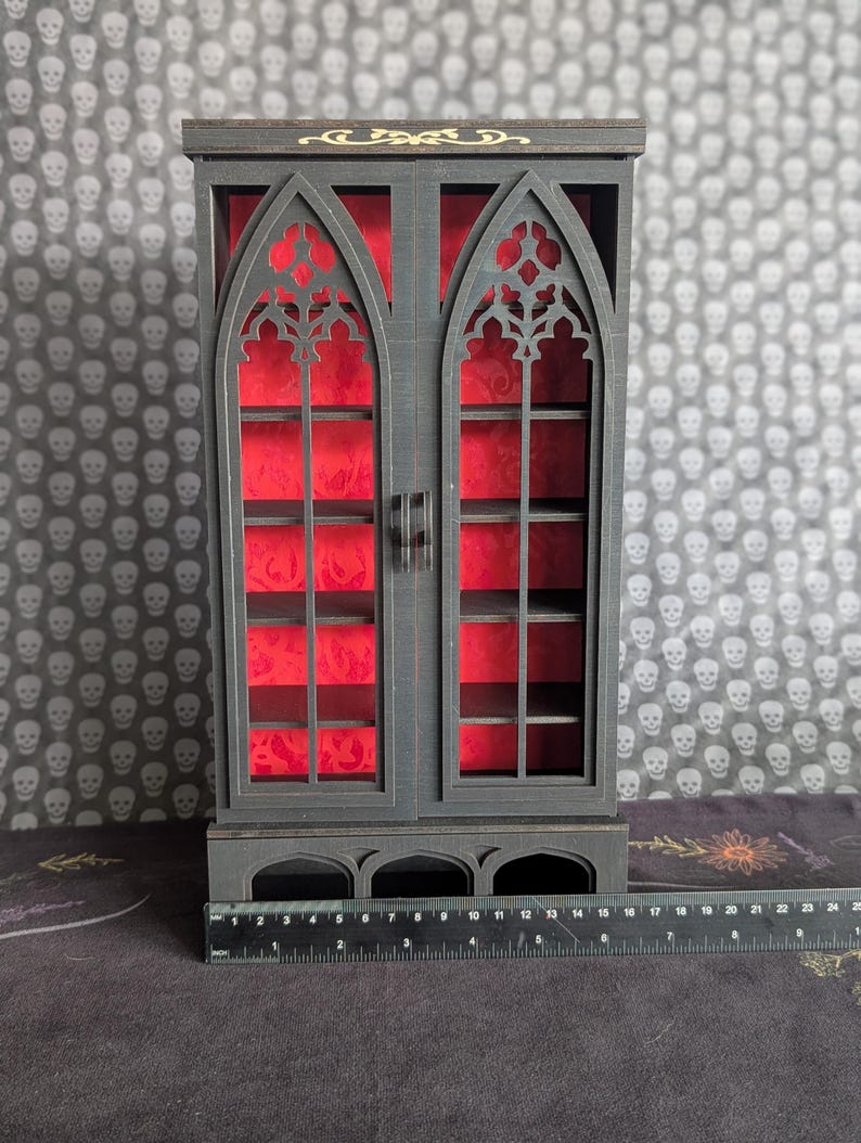 K&ouml;nnte beinhalten: Ein schwarzer Holzschrank mit zwei T&uuml;ren, der gotische Bogenfenster-Designs aufweist. Der Innenraum ist mit rotem Stoff ausgekleidet. Der Schrank hat Regale und ist etwa 25 cm breit.