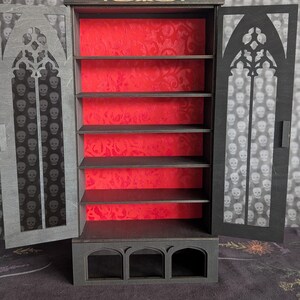 K&ouml;nnte beinhalten: Ein schwarzer Holzschrank im gotischen Stil mit roter Innenausstattung und mehreren Regalen. Die Schrankt&uuml;ren haben ein dekoratives Ausschnittmuster mit Totenk&ouml;pfen. Der Schrank ist ge&ouml;ffnet und gibt die Regale frei.