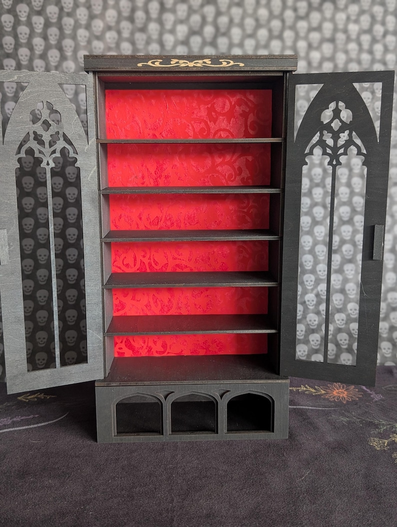 K&ouml;nnte beinhalten: Ein schwarzer Holzschrank im gotischen Stil mit roter Innenausstattung und mehreren Regalen. Die Schrankt&uuml;ren haben ein dekoratives Ausschnittmuster mit Totenk&ouml;pfen. Der Schrank ist ge&ouml;ffnet und gibt die Regale frei.