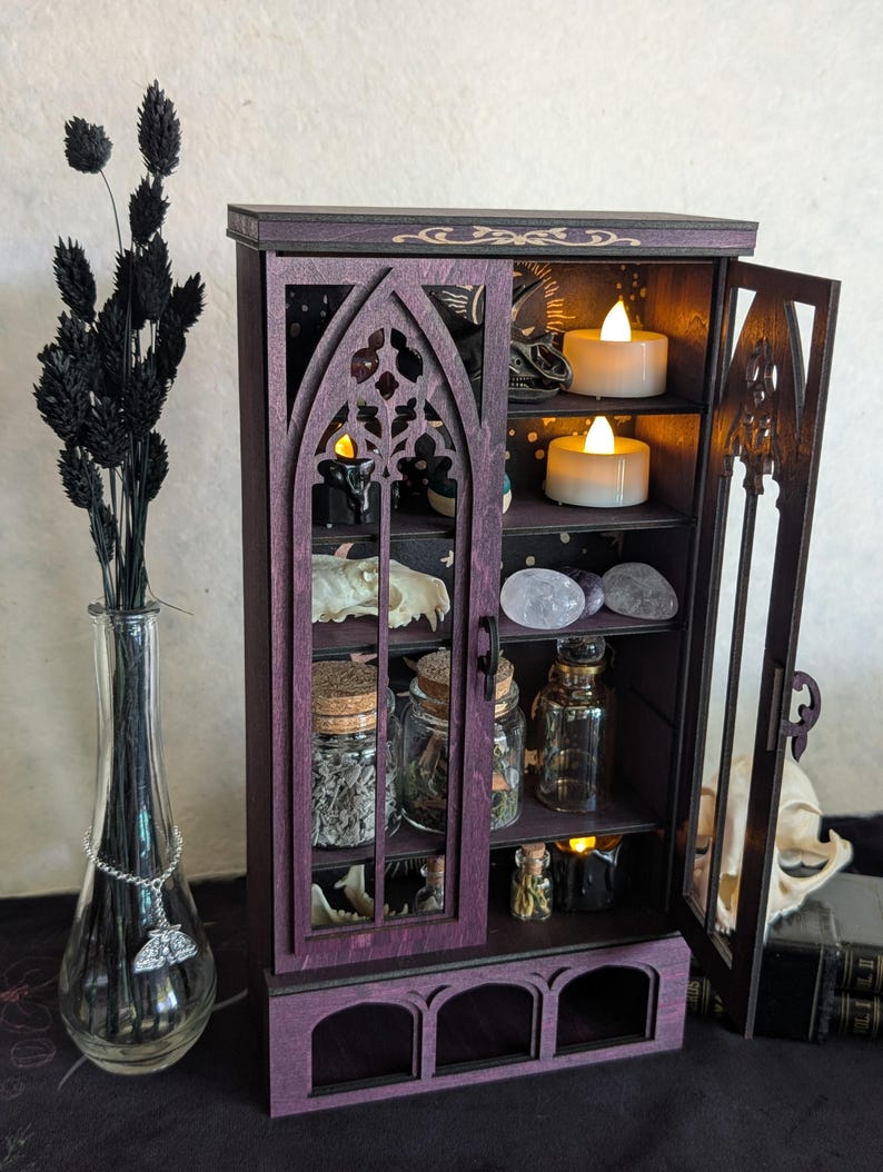 Puede incluir: Un armario de madera morada con puertas de arco g&oacute;tico y varios estantes que contienen frascos de vidrio, cristales, cr&aacute;neos de animales y peque&ntilde;as velas LED. Junto al armario hay un jarr&oacute;n de vidrio transparente con tallos negros.