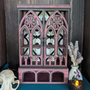 May include: A miniature wooden cabinet with a gothic design, painted in shades of pink and gray. The cabinet features arched doors with decorative cutouts and interior shelves. A small skull, vase with dried flowers, and a candle are in the foreground.
