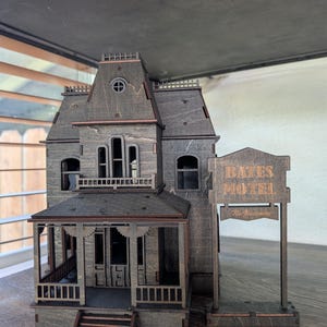 May include: A wooden model of the Bates Motel, featuring a detailed facade with a porch, windows, and a sign that reads "Bates Motel" and "No Vacancy". The model is made of dark wood and has intricate cut-out details.