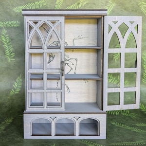 May include: A light gray wooden cabinet with an open door and decorative cutouts. The cabinet has shelves and a base with arched openings. The interior features a skeletal horse illustration. The cabinet is on a green surface.