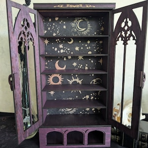 Puede incluir: Un gabinete de madera de color morado oscuro con dos puertas abiertas que presentan recortes de arco g&oacute;tico. El panel trasero interior es negro con patrones celestiales dorados de lunas, soles y estrellas. Contiene cinco estantes y tres compartimentos.