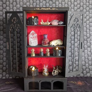 K&ouml;nnte beinhalten: Ein schwarzer Holzschrank mit T&uuml;ren und Regalen im gotischen Stil, der verschiedene Gegenst&auml;nde ausstellt. Das Innere des Schranks ist rot und enth&auml;lt Kerzen, Kristalle, kleine Gl&auml;ser und dekorative Sch&auml;del. Der Hintergrund zeigt ein Totenkopfmuster.