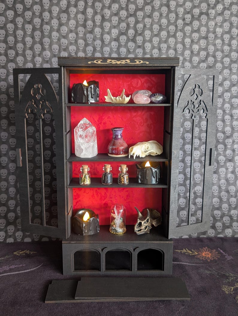 K&ouml;nnte beinhalten: Ein schwarzer Holzschrank mit T&uuml;ren und Regalen im gotischen Stil, der verschiedene Gegenst&auml;nde ausstellt. Das Innere des Schranks ist rot und enth&auml;lt Kerzen, Kristalle, kleine Gl&auml;ser und dekorative Sch&auml;del. Der Hintergrund zeigt ein Totenkopfmuster.