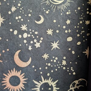 Puede incluir: Un papel texturizado de color gris carb&oacute;n oscuro con un patr&oacute;n repetitivo de elementos celestiales en oro met&aacute;lico, que incluye soles con rayos, lunas crecientes, varias formas de estrellas y peque&ntilde;os puntos circulares.