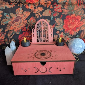 May include: A pink wooden altar box with a decorative arched cutout, moon phase cutouts, and a celestial design. It features two LED candles, clear quartz crystals, and a crystal ball on a gold stand. The background is a floral pattern.