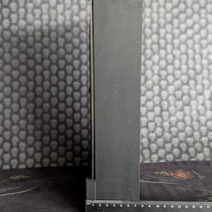 K&ouml;nnte beinhalten: Ein schwarzes, rechteckiges Objekt mit einem Lineal daneben. Das Lineal zeigt Messungen in Zentimetern an. Das Objekt befindet sich vor einem Hintergrund mit einem sich wiederholenden Totenkopfmuster.
