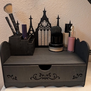 May include: A black wooden makeup organizer with a drawer and multiple compartments. The organizer features a gothic arch design and holds makeup brushes, lipsticks, and bottles. The drawer has a decorative floral design.