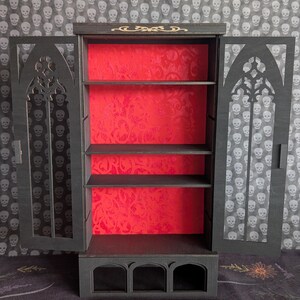 K&ouml;nnte beinhalten: Ein schwarzer Holzschrank mit gotischen Bogen-T&uuml;ren. Der Innenraum hat eine rote, gemusterte R&uuml;ckwand und drei Regale. Der Schrank ist ge&ouml;ffnet und zeigt die Regale. Der Hintergrund ist ein Totenkopfmuster.