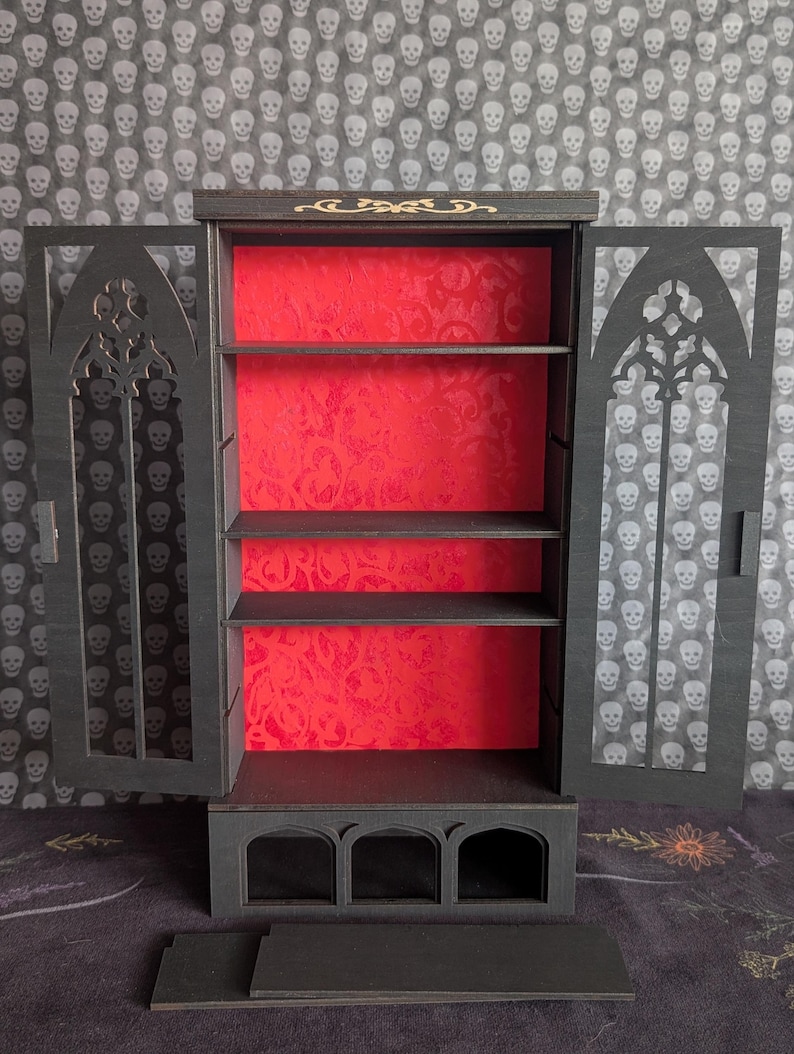 K&ouml;nnte beinhalten: Ein schwarzer Holzschrank mit gotischen Bogen-T&uuml;ren. Der Innenraum hat eine rote, gemusterte R&uuml;ckwand und drei Regale. Der Schrank ist ge&ouml;ffnet und zeigt die Regale. Der Hintergrund ist ein Totenkopfmuster.
