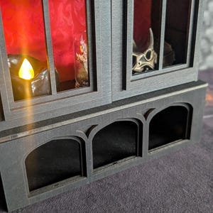 K&ouml;nnte beinhalten: Ein dunkelgrauer Holzschrank mit Glast&uuml;ren und bogenf&ouml;rmigen Ausschnitten. Der Innenraum des Schranks hat einen roten Hintergrund und enth&auml;lt kleine Glasfl&auml;schchen und einen Sch&auml;del. Eine brennende Kerze ist sichtbar.