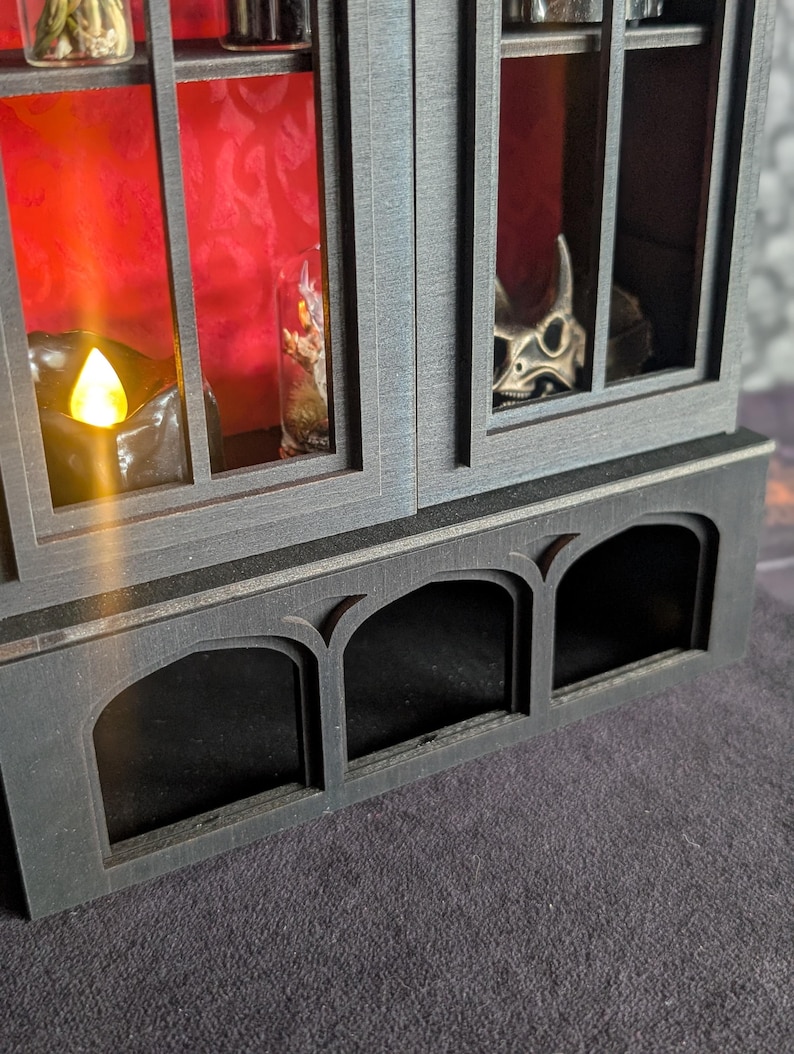 K&ouml;nnte beinhalten: Ein dunkelgrauer Holzschrank mit Glast&uuml;ren und bogenf&ouml;rmigen Ausschnitten. Der Innenraum des Schranks hat einen roten Hintergrund und enth&auml;lt kleine Glasfl&auml;schchen und einen Sch&auml;del. Eine brennende Kerze ist sichtbar.