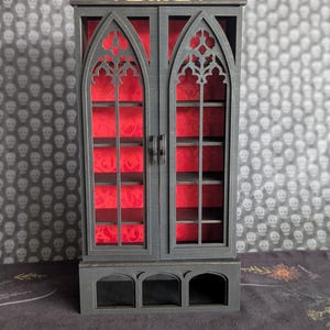 K&ouml;nnte beinhalten: Ein kleiner schwarzer Schrank mit gotischem Bogen-Design. Der Schrank hat zwei T&uuml;ren mit aufwendigen Ausschnitten, die einen roten Innenraum zeigen. Er hat Regale und einen dekorativen Sockel. Der Hintergrund hat ein Totenkopfmuster.