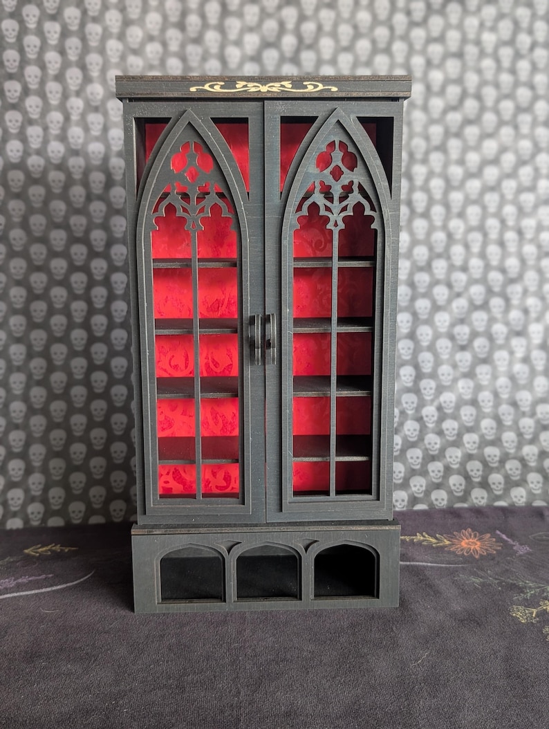 K&ouml;nnte beinhalten: Ein kleiner schwarzer Schrank mit gotischem Bogen-Design. Der Schrank hat zwei T&uuml;ren mit aufwendigen Ausschnitten, die einen roten Innenraum zeigen. Er hat Regale und einen dekorativen Sockel. Der Hintergrund hat ein Totenkopfmuster.