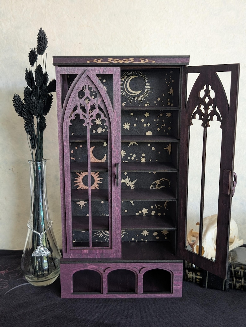 Puede incluir: Un armario de madera morada con dos puertas abatibles que presentan recortes arqueados de estilo g&oacute;tico. El panel trasero interior es negro con motivos celestiales dorados, incluyendo lunas, soles y estrellas. La base incluye tres peque&ntilde;os compartimentos abiertos.