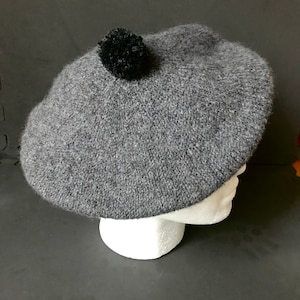 May include: A gray knit beret with a black pom-pom on top. The hat is displayed on a white foam head form against a dark background. The beret is a classic style, suitable for various occasions.