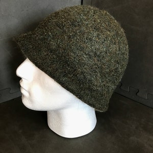 May include: A dark green cloche hat with a short brim. The hat is made of a textured, fuzzy material. The hat is displayed on a white mannequin head against a dark background.