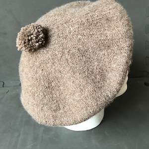 May include: A beige beret with a small pom-pom on the side. The hat is made of a textured, knitted material. The beret is displayed on a white mannequin head.