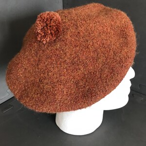 May include: A rust-colored beret with a textured wool appearance. A small, matching pom-pom is attached to the side. The beret is displayed on a white mannequin head. This hat is a stylish accessory.