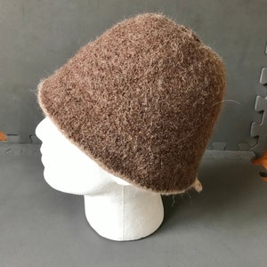 Könnte beinhalten: Ein brauner, strukturierter Hut mit einer abgerundeten Krone und einem leicht ausgestellten Rand. Der Hut wird auf einem weißen Schaufensterkopf präsentiert, der seine Form und Textur zeigt. Der Hut scheint aus einem wollähnlichen Material zu bestehen.