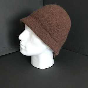 May include: A brown cloche hat with a small, downward-sloping brim. The hat is made from a textured, fuzzy fabric and is shown on a white mannequin head. The hat's colour is a warm, earthy brown, ideal for cooler temperatures.