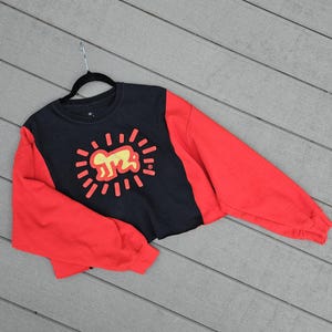 Puede incluir: Sudadera corta negra y roja con un gr&aacute;fico amarillo de una persona rodeada de l&iacute;neas radiantes. La sudadera tiene mangas largas rojas y un torso negro con el gr&aacute;fico en el centro.