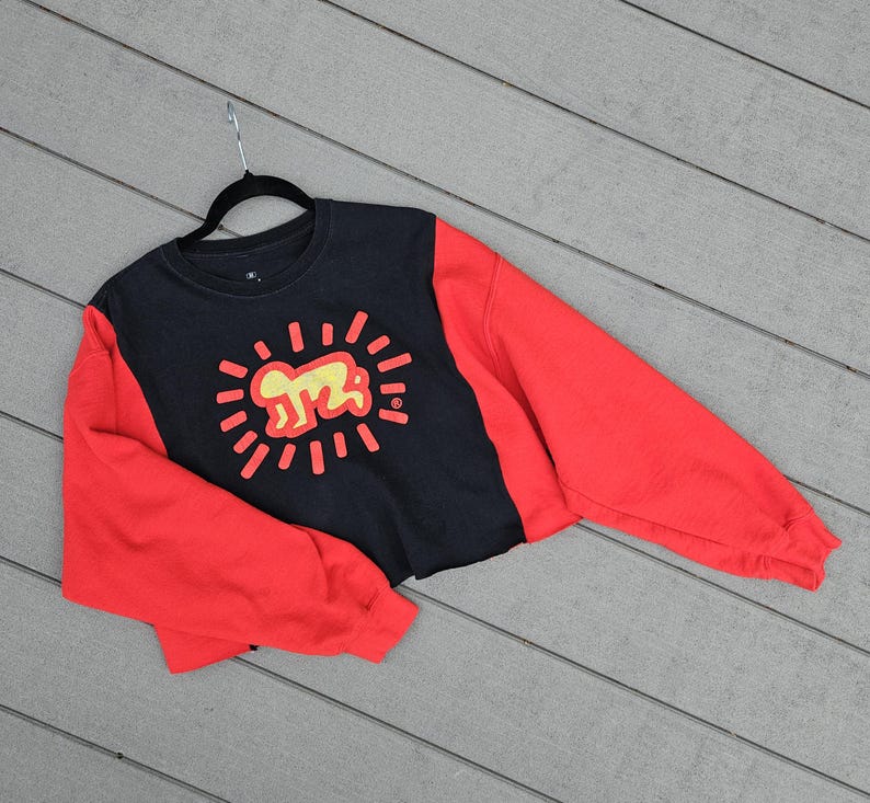 Puede incluir: Sudadera corta negra y roja con un gr&aacute;fico amarillo de una persona rodeada de l&iacute;neas radiantes. La sudadera tiene mangas largas rojas y un torso negro con el gr&aacute;fico en el centro.