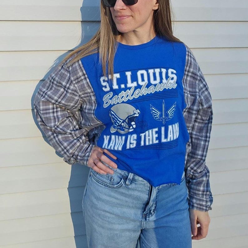 Pu&ograve; includere: Una maglia a maniche lunghe con pannello centrale blu reale e maniche a quadri grigi. Il davanti presenta la scritta bianca ST. LOUIS BATTLEHAWKS, un casco da football, il logo di una spada e la scritta KAW IS THE LAW in lettere bianche.