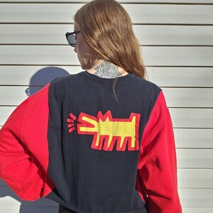 Puede incluir: Una sudadera recortada con un dise&ntilde;o de bloques de color. Las mangas son rojas y el cuerpo es negro con un gr&aacute;fico de perro amarillo y rojo en la espalda. La sudadera es usada por una persona.